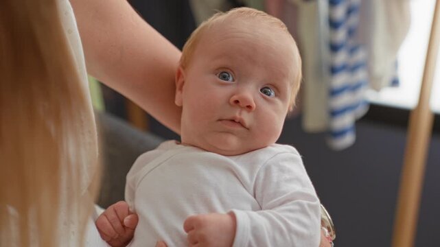 Baby held by mother, face and bare cheek gazing to the side, blue eyes focused, wearing white onesie in a building; quiet curiosity.