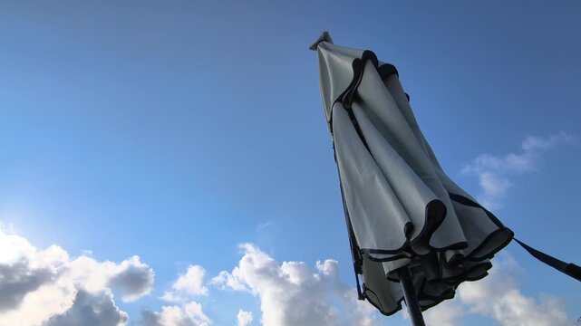 Low angle view of a closed parasol waving in the wind against a blue sky with fluffy clouds passing by, representing the end of summer vacation and the coming storm