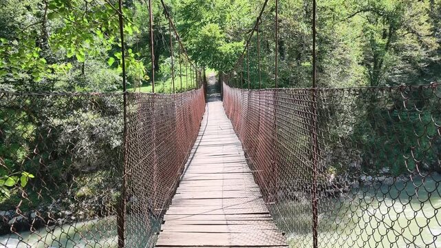 Crossing narrow old wooden suspension bridge over a mountain river in a lush green forest, representing adventure, exploration, risk, and the journey forward