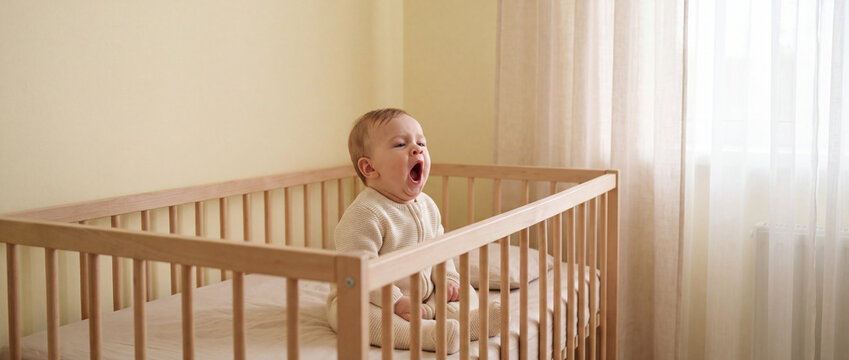 A curious baby stands inside a wooden crib in a sunlit nursery room looking upwards. peaceful morning routine, safe child environment, happy nursery lifestyle, innocent home moment.
