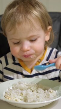 Cute toddler girl learning to eat rice by himself at home. Close up of caucasian baby holding a blue spoon and eating. User Generated Content. Vertical B-roll for parenting and baby food concept.