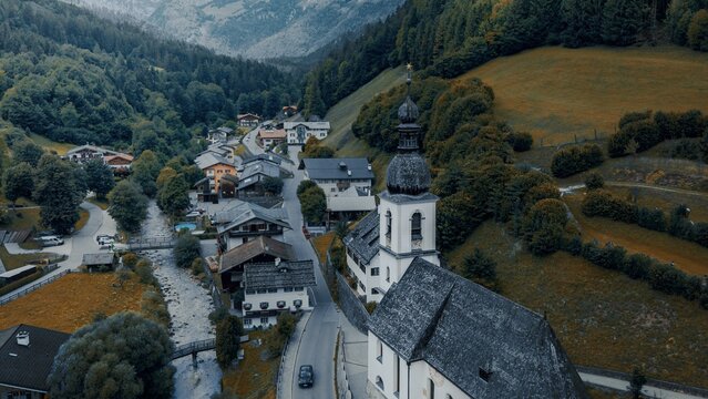 Aerial view of the Parish Church of St. Sebastian surrounded by traditional alpine houses, a flowing river, and lush green mountain slopes, Ramsau bei Berchtesgaden, Bavaria, Germany.