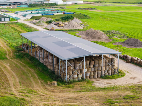Aerial view of a large agricultural shed filled with stacked round hay bales in a rural landscape with green fields and dirt paths in Virpe, Ventspils novads, Latvia.