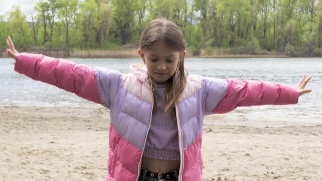 Little girl in puffer jacket playing and gesturing by the river on a spring day