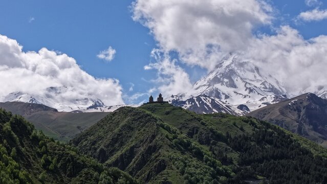 Aerial view of Gergeti Trinity Church perched on a green hill against the backdrop of snow-capped Mount Kazbek under a blue sky Stepantsminda, Mtskheta-Mtianeti, Georgia.