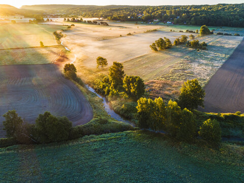 Aerial view of the rural landscape in Hallesaker featuring a winding stream, agricultural fields, and lush trees illuminated by golden hour light in Hallesaker, Halland County, Sweden.