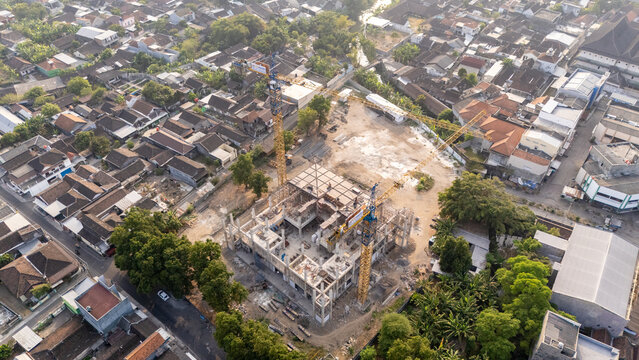 Aerial view of a construction site with two tall yellow cranes, concrete building frame, and surrounding residential neighborhood in Surakarta, Central Java, Indonesia.