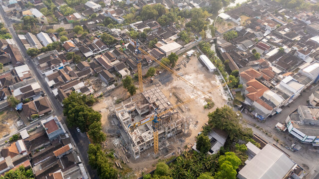 Aerial view of a construction site with tower cranes and building foundations surrounded by dense residential rooftops and lush trees in Surakarta, Central Java, Indonesia.