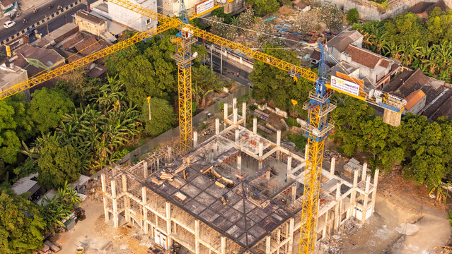 Aerial view of a construction site featuring two yellow tower cranes and a concrete building frame surrounded by lush trees and urban roads in Surakarta, Central Java, Indonesia.