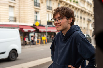 A young man wearing eyeglasses and a blue hoodie stands on a city street in Paris, France. Traditional European architecture and storefronts are visible in the background. © dsbedarieva