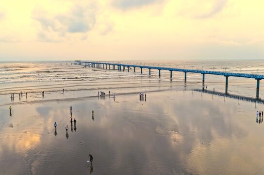 Aerial view of the BD Navy Jetty extending into the sea at low tide with people walking on the reflective wet sand in Cox's Bazar District, Chittagong Division, Bangladesh.