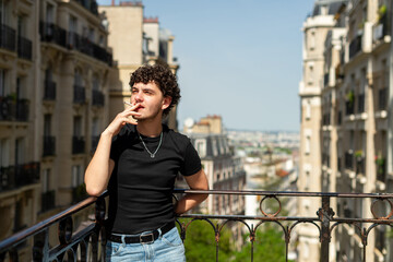 A young man stands leaning on a metal balcony railing smoking a cigarette in Paris, France. Traditional residential buildings frame the background view of the city. © dsbedarieva