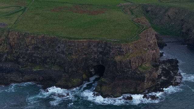 Aerial view of the Giant's Causeway coastline, featuring dramatic basalt cliffs, a sea cave, and green grassy clifftops meeting the Atlantic Ocean in Bushmills, Northern Ireland, United Kingdom.