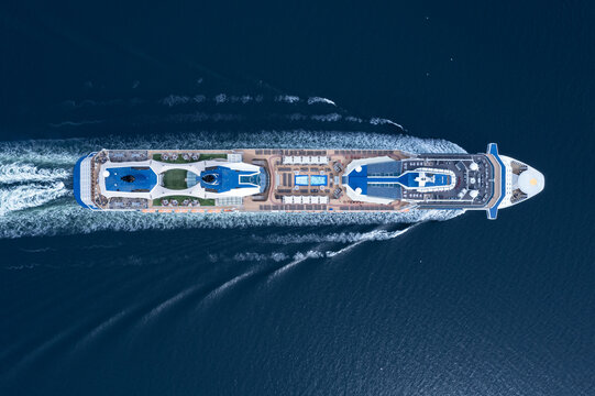 Bute, Scotland - 27 July 2022: Aerial view of a large cruise ship sailing through deep blue waters, leaving a white wake behind it, with visible decks, swimming pools, and sun loungers.