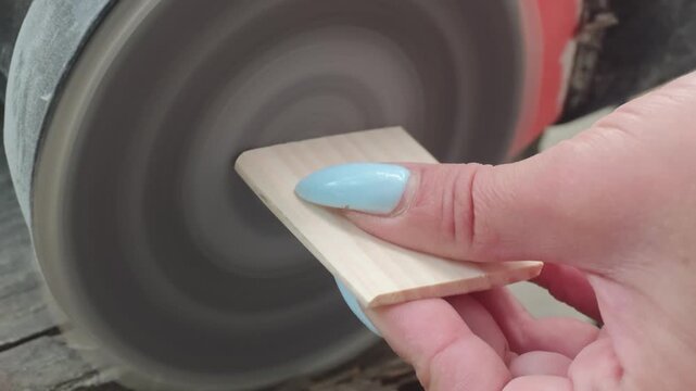 Close-up of a woman with blue manicured nails using a disc sanding machine to smooth and round the edges of a small wooden plank in a carpenter workshop during woodworking process.