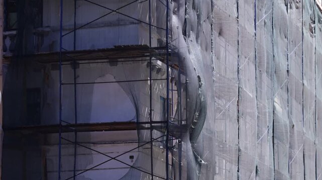 Urban scene shows building facade with scaffolding and protective netting; visible structural openings and weathered surfaces convey active renovation mood