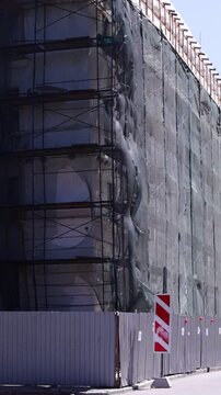 Urban scene shows building facade with scaffolding and protective netting; visible structural openings and weathered surfaces convey active renovation mood