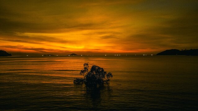 Painan, Sumatera Barat, Indonesia - 22 March 2022: Aerial view of a small tree island in the calm sea during a vibrant golden sunset.