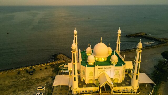 Padang, Indonesia - 22 March 2022: Aerial view of Masjid Al Hakim mosque situated on the coast with its white domes and minarets illuminated against the calm sea at sunset.