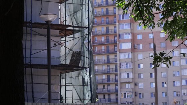 Urban scene shows building facade with scaffolding and protective netting; visible structural openings and weathered surfaces convey active renovation mood
