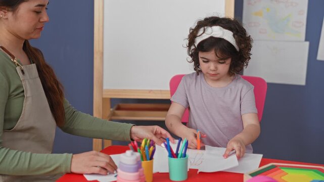 Woman guiding girl's hand while cutting paper with scissors at art studio; early learning concentration.