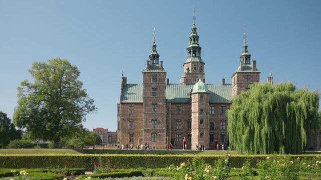 Historic Rosenborg Castle stands in the royal park in Copenhagen, Denmark. Lush green trees frame the renaissance brick palace. Tourists explore the famous Danish landmark on a sunny summer day.