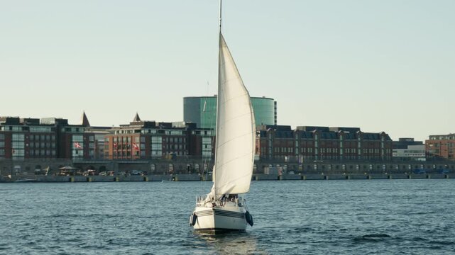 Sailboat navigates calm harbor water in Copenhagen. Modern urban waterfront lines scenic Nordic canal during sunny summer voyage. Recreational yacht cruising offers active tourism and leisure.