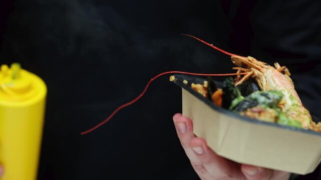 Man holding takeout box with fried rice, whole prawn and mussels with green sauce. Street food meal in plastic container. Seafood take away dish concept.