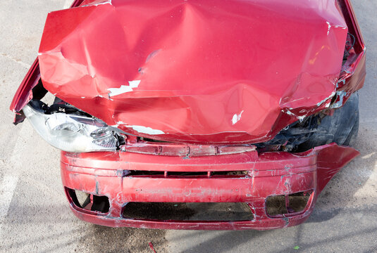 Damaged red sport compact car after a severe traffic accident on a city street side. Smashed front bumper, crumpled bonnet hood, broken headlight optics, and crushed fender panel. Auto insurance. 