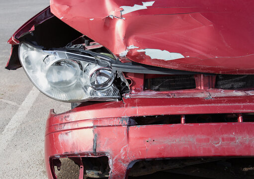 Damaged red sport compact car after a severe traffic accident on a city street side. Smashed front bumper, crumpled bonnet hood, broken headlight optics, and crushed fender panel. Auto insurance. 