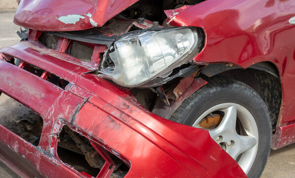 Damaged red sport compact car after a severe traffic accident on a city street side. Smashed front bumper, crumpled bonnet hood, broken headlight optics, and crushed fender panel. Auto insurance. 