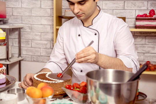 Pastry chef decorating a chocolate cake with fresh strawberries and blueberries using tongs in a confectionery workshop