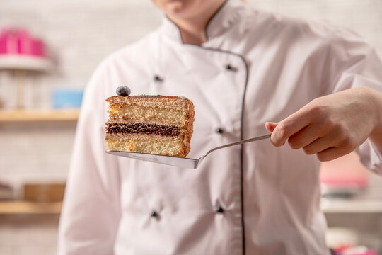 Hand holding a slice of layered chocolate sponge cake on a server, with decorated desserts in a pastry workshop.