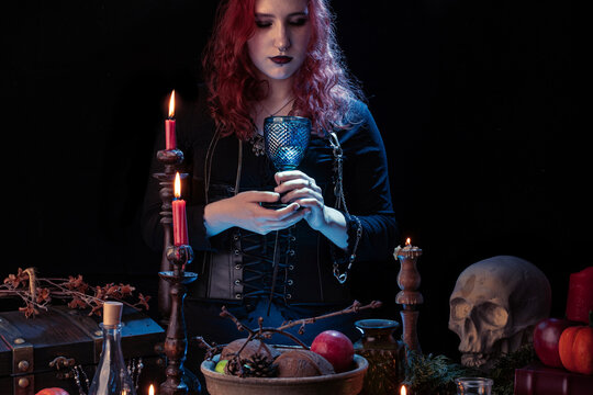 Witch conjuring a love potion, cooking up love, casts a spell glowing poison. Young woman, a magician girl with red hair, dressed in Halloween attire.