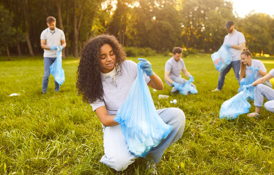 Happy young people volunteers with trash bags collecting garbage from green grass in summer park. Men and women cleaning trash in nature. Environmental pollution, volunteering and ecology concept