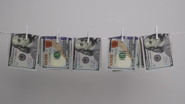 Five US hundred dollar banknotes hang folded in half with wooden clothespins on a white rope, showing money laundering.