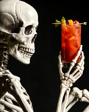 Halloween red cocktail on black background. Skeleton is drinking alcohol at the counter. Alcoholic drink on dark stone table. Spooky, scary, fun, horror, creative. Bar holiday menu. Vertical