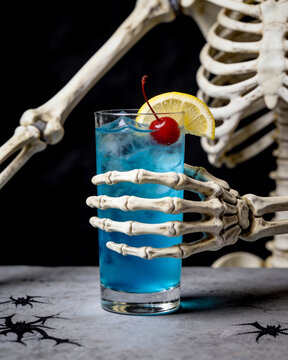 Halloween blue lagoon cocktail on black background. Skeleton is drinking alcohol at the counter. Alcoholic drink on dark stone table. Spooky, scary, fun, horror, creative. Bar holiday menu. Vertical