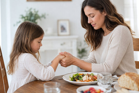 Mother and daughter holding hands and praying before healthy dinner at home, concept of faith, gratitude, parenting, and teaching religious family values in daily life.