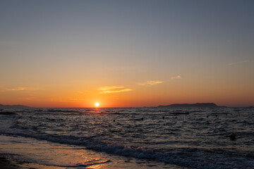 Obraz premium Sun touches sea horizon casting orange glow over wavy dark water. Coastal view with wet sandy beach shore, light clouds in sky, and distant island silhouette.