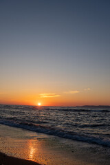 Obraz premium Sun touches sea horizon casting orange light on rippling waves. Coastal view with wet beach shore, sunset reflection on sand, gradient sky, and distant island hills.