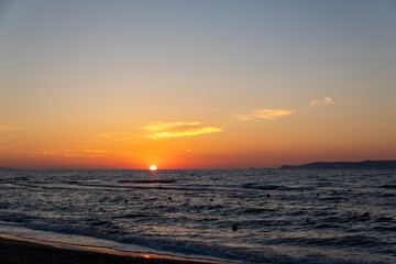 Obraz premium Sun touches sea horizon casting orange light on dark wavy water. Coastline view with sandy beach shore, small clouds in twilight sky, and distant island silhouette.