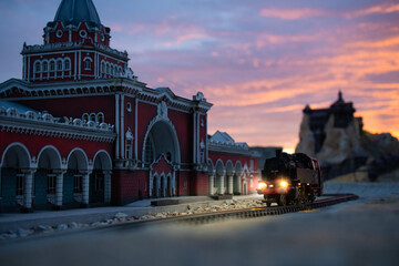 Miniature steam locomotive with glowing headlights traveling past a red railway station at dusk © ArtExpert