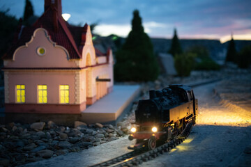 Miniature steam locomotive passing an illuminated pink railway station at dusk © ArtExpert