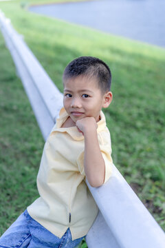 A charming portrait of a cute young Asian boy leaning on a white metal guardrail in a scenic park, capturing a sweet family lifestyle moment with warm natural lighting and a calm, gentle expression.