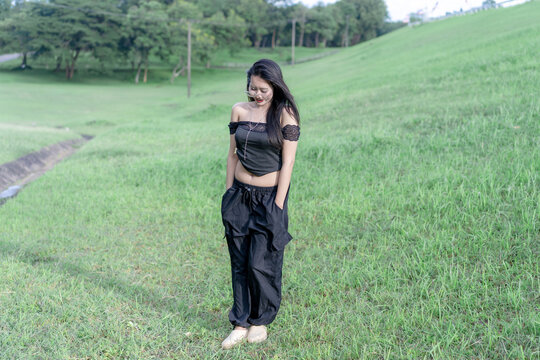 A thoughtful young Asian woman standing on a lush green hill by a serene reservoir, looking down with her hands in pocket while wearing glasses and a stylish black outfit on a bright sunny summer day.