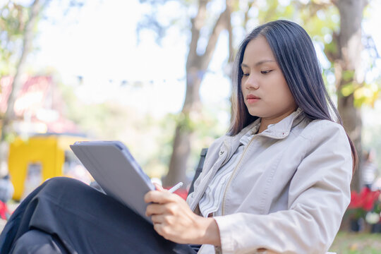 A focused young Asian woman sitting outdoors in a scenic park, working and writing on a digital tablet with a stylus pen while wearing a light jacket and casual shirt surrounded by lush green foliage.