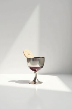 A solitary silver goblet, partially filled with a dark liquid, rests on a pristine white surface, a single round cracker delicately perched on its rim, bathed in soft, natural light.