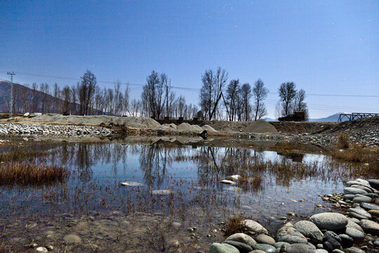 View of a calm pond reflecting bare trees under a clear blue sky, with piles of gravel and smooth river stones in the foreground in Khwazakhela, Khyber Pakhtunkhwa, Pakistan.