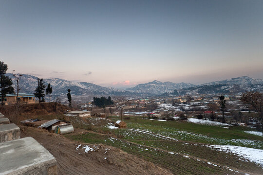 View of the snowy landscape of Chattar Plain with mountains in the background, green fields with patches of snow, and rural buildings in Chattar Plain, Khyber Pakhtunkhwa, Pakistan.